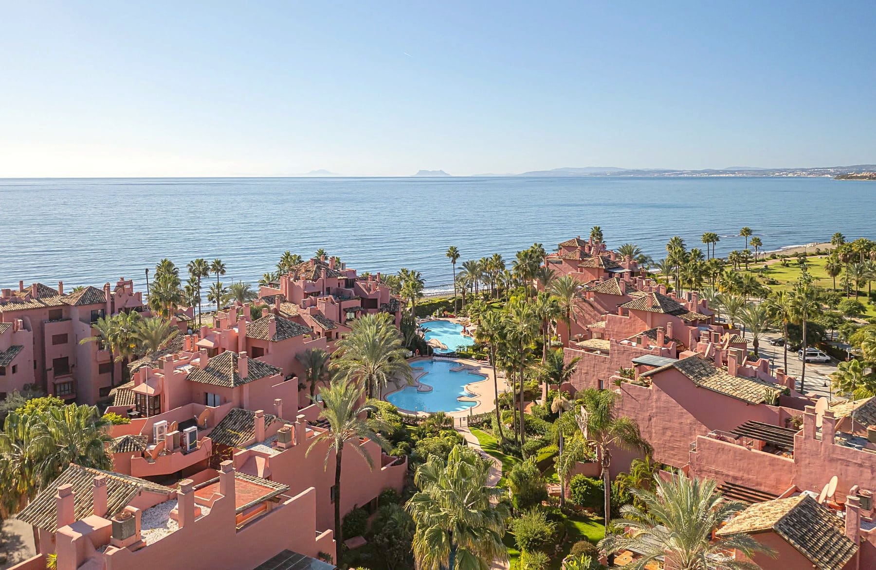 Terracotta buildings with pools and palm trees by the sea, with Gibraltar on the horizon